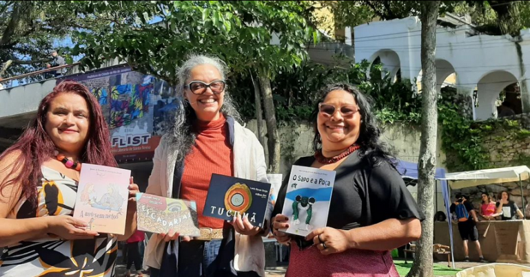 Flist 2022, Pré-lançamento dos livros de Maria do Rosário (Duaia) de Assumpção Braga pela Realizamos Artes no Parque das Ruínas, RJ. Em área aberta, ao sol, há céu azul, árvores e ao fundo na parte superior direita, a escadaria coberta do Parque das Ruínas. Ao fundo na parte direita inferior, há pessoas e um toldo, em movimento de feira. À frente, mais para a esquerda, Arlene Costa, Duaia Assumpção e Lucia Moraes Tucuju sorriem para a câmera exibindo livros. Arlene é morena clara, tem cabelo avermelhado, usa vestido estampado e um colar vermelho e preto. Mostra o livro “A Avó sem netos” de Duaia. Duaia mostra o livro “Escadarias”, de Arlene e o livro “Tucumã”, de Lucia Moraes Tucuju. Duaia é morena clara, também sorri para a câmera, usa óculos com lentes levemente escuras, moldura preta com uma barra branca na parte superior, blazer creme, sobre blusa vermelha e calça azul escura com cinto de estampa de tigre. Seu cabelo longo e grisalho tem uma mecha e está semi preso. Lucia Moraes Tucuju é indígena, tem cabelos pretos longos e lisos, usa óculos escuros, colar vermelho e preto, anel, blusa preta e saia vermelha. Lucia mostra o livro “O Sapo e a Poça” de Duaia.
