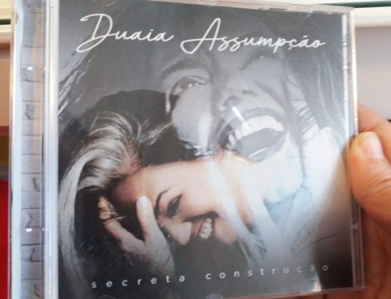 Mão segura a Capa do CD Secreta Construção, de Duaia Assumpção. Ao clicar, o link encaminha para a página da cantora em seu site.