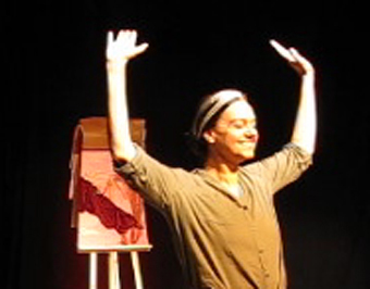 Duaia Assumpção, braços levantados, sorri ao final do espetáculo 4 Peça de Câmara para 1 Atriz e 4 Personagens. Ao fundo, um painel colorido sobre cavalete.