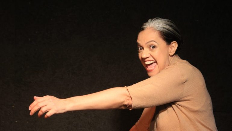 4 – Peça de Câmara para 1 Atriz e 4 Personagens. Sobre fundo negro, à direita, Duaia Assumpção de perfil olha a câmera com grito em “A”, braço esquerdo estendido em meio a movimento. Foto de Claudia Ribeiro.