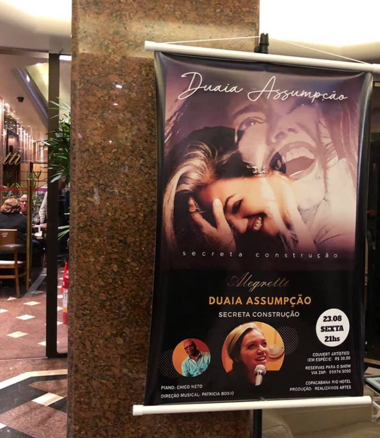 Banner na porta do bar Alegretti, com foto da capa do CD de Duaia Assumpção, Secreta Construção. 23.08, sexta 21hs. Piano: Chico Neto. Direção Musical: Patricia Bosio. Foto de Chiquinho e de Duaia. (Couvert Artístico em espécie R$ 30,00). Reservas para o show via zap (cel). Copacabana Rio Hotel. Produção: Realizamos Artes.